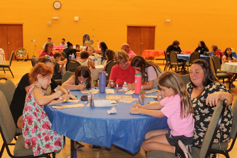 Adults and children sit around a blue-covered table in the South Cape Community Center gym, concentrating on their bingo cards during a family bingo event.